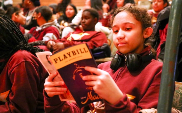 A young student wearing a red sweatshirt sits in a packed auditorium and examines her Playbill