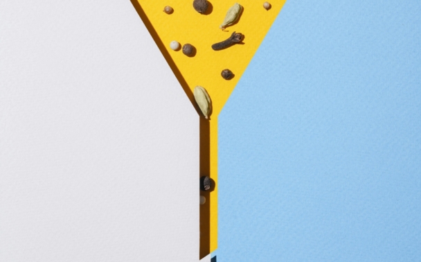 A yellow martini glass shape on a split white and blue background, filled with assorted spices and a garlic clove, with a single drop of liquid falling from the glass stem.