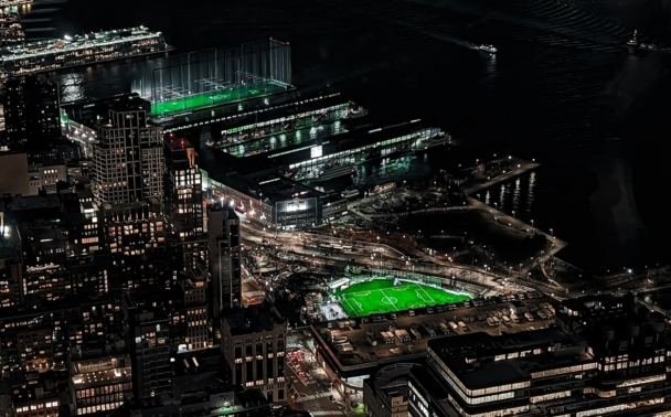 A nighttime aerial view of a city with illuminated buildings and streets, featuring two bright green-lit sports fields near a waterfront, with reflections of city lights on the water.