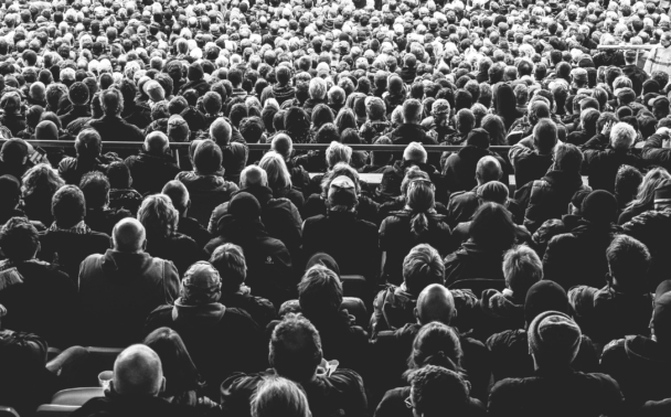 A large crowd of people seen from behind, all seated and facing forward. The black and white photo captures the dense audience in an outdoor or indoor venue, with heads and shoulders filling the frame.