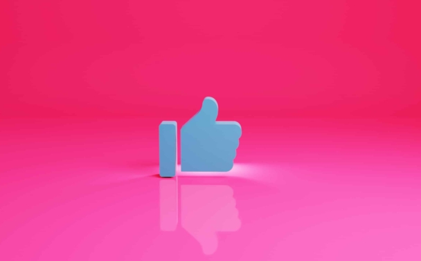 A blue thumbs-up icon stands upright on a glossy pink surface with a bright pink background, creating a simple and modern composition.