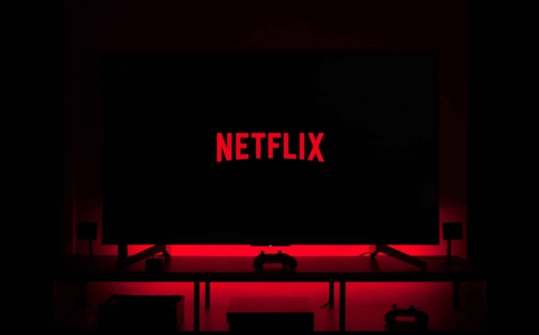 A dark room with a TV displaying the red Netflix logo on a black screen, illuminated by red backlighting beneath the TV stand, with gaming controllers and speakers visible nearby.