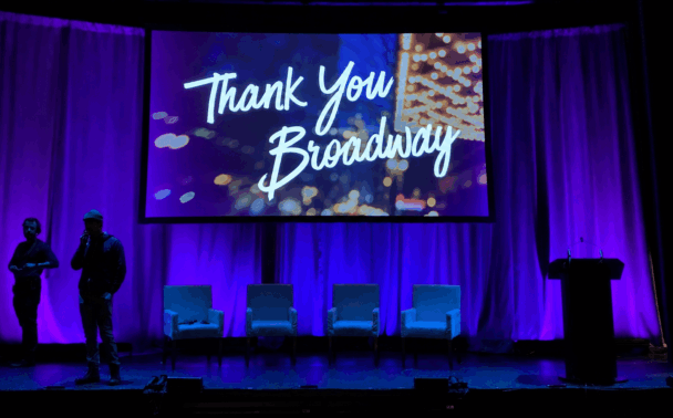 A stage with four empty chairs, a podium, two people standing, and a large screen displaying Thank You Broadway in white script, all illuminated by purple lighting.