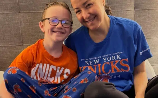 A smiling woman and child sit close together on a bed, both wearing New York Knicks shirts. The child wears glasses and has their arm around the woman. The background shows a gray upholstered headboard.