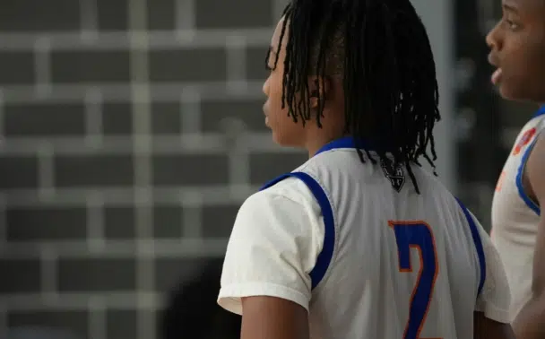 A basketball player wearing a white jersey with blue accents and the number 2 on the back stands on the court, facing away from the camera, with another player visible beside them. Brick wall is in the background.