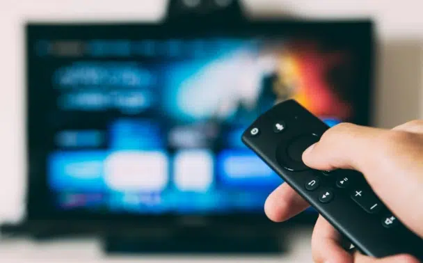 A hand holding a TV remote control, pointed toward a television screen displaying a blurred, colorful user interface, suggesting streaming or browsing content.