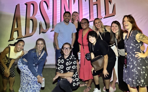 A group of eleven people smile and pose in front of a large white structure with ABSINTHE written in bold letters, illuminated by colorful lights at night.