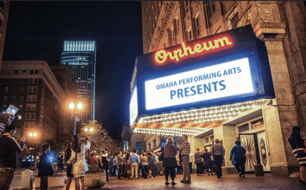 People gather outside the lit-up Orpheum Theatre at night. The marquee reads “Omaha Performing Arts Presents.” City buildings and glowing streetlights are visible in the background.