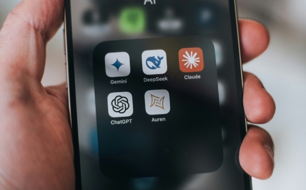 A hand holds a smartphone displaying an AI folder with five app icons: Gemini, DeepSeek, Claude, ChatGPT, and Auren. The background is blurred.