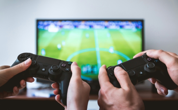 Two people hold video game controllers, playing a soccer game on a large TV screen in a living room setting.