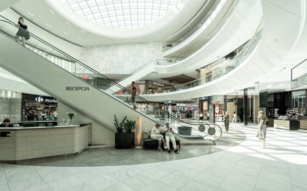 A bright, modern shopping mall interior with curved balconies, an escalator, stores, seated people, and a glass dome ceiling allowing natural light inside. Shoppers walk and interact throughout the space.