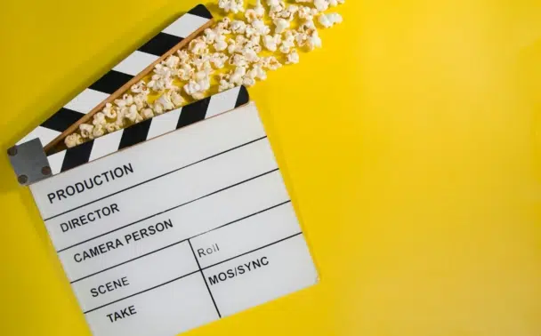 A film clapperboard with popcorn spilling out lies on a bright yellow background, suggesting a movie or film theme.