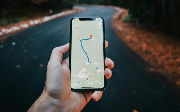 A person holds a smartphone with a map and route displayed, standing on a winding road surrounded by autumn trees.