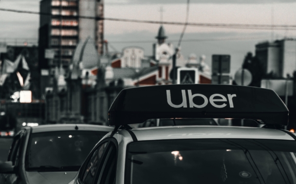A white Hyundai car with an Uber sign on the roof is driving on a city street at dusk, followed by other vehicles. Buildings and streetlights are visible in the background.