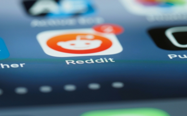 A close-up, shallow depth-of-field shot of a smartphone home screen, focusing on the Reddit app icon, which shows a notification badge. Other app icons are visible but blurred in the background.