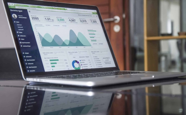 A laptop displaying a data analytics dashboard with graphs, charts, and numerical statistics sits on a reflective desk in an office setting.