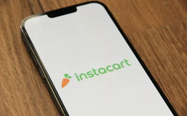 A smartphone with the Instacart app open displays the Instacart logo—a carrot icon and the text instacart—on a wooden surface.