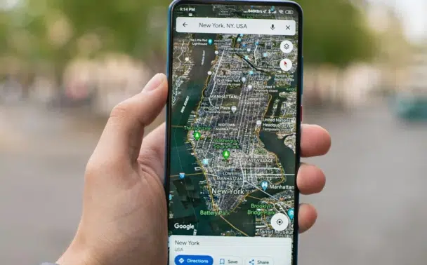 A hand holds a smartphone displaying a satellite map view of Manhattan, New York on Google Maps.